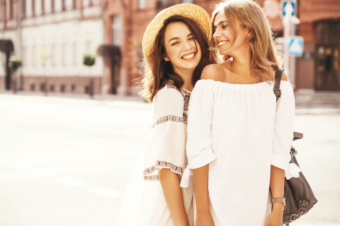 Fashion portrait of two young stylish hippie brunette and blond women models in summer sunny day in white hipster clothes posing on the street background. No makeup